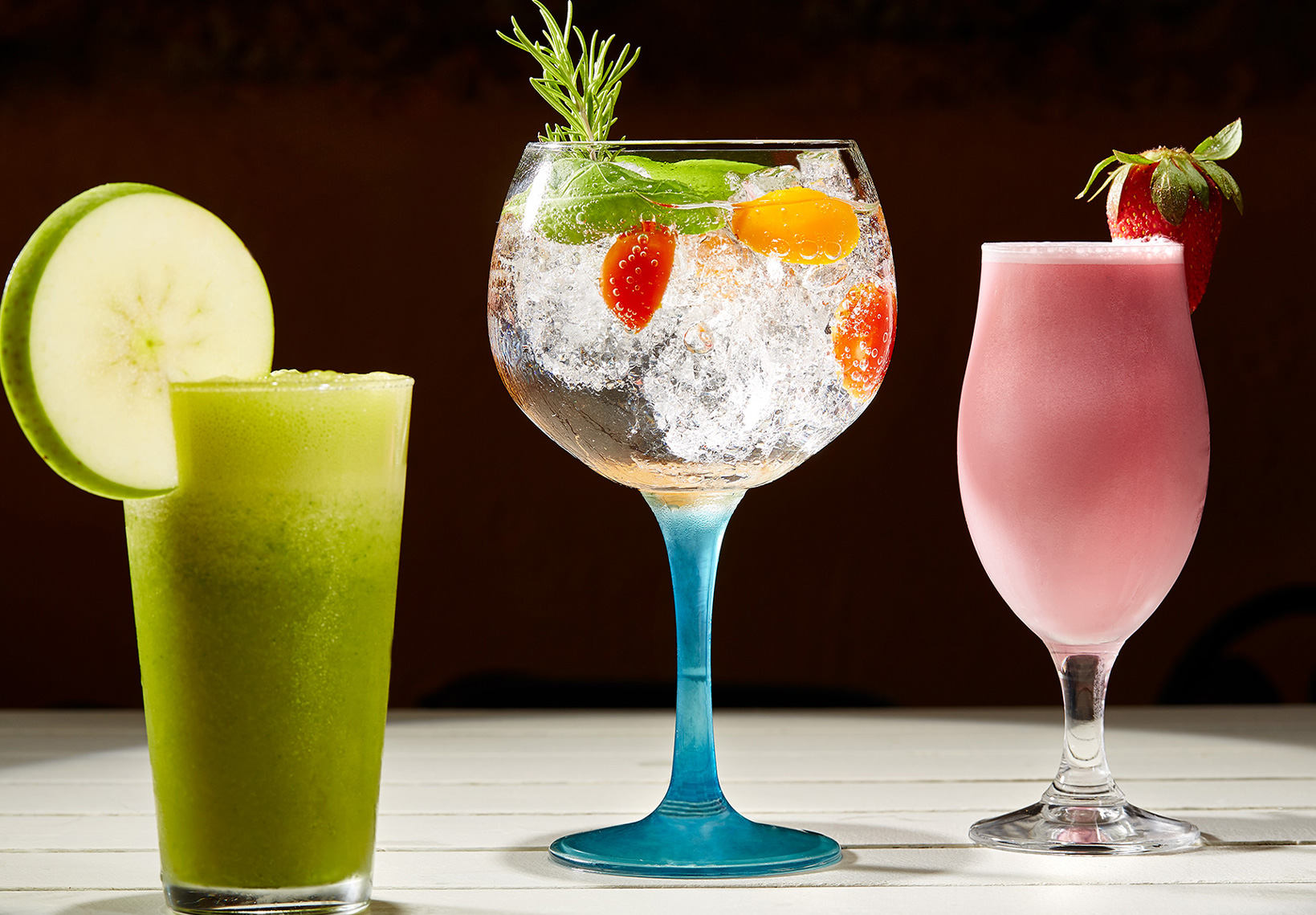 Três cocktails coloridos (verde, transparente com tomate e rosa) alinhados sobre uma mesa branca.