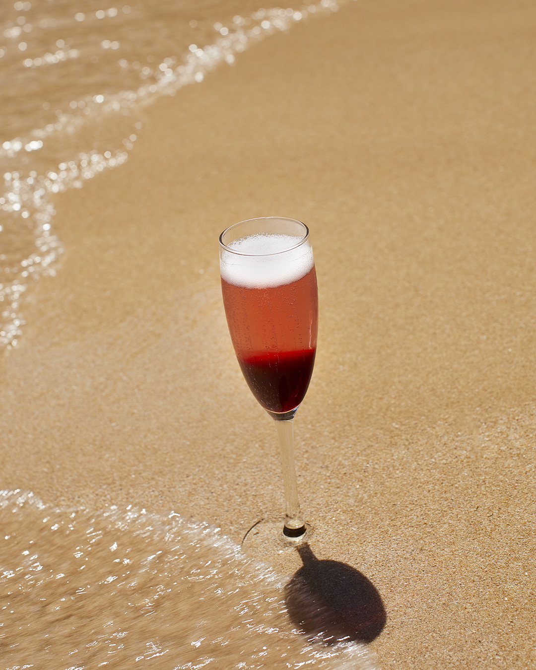 Uma flute de espumante rosado com espuma branca, espetada na areia húmida da praia enquanto uma onda se aproxima.