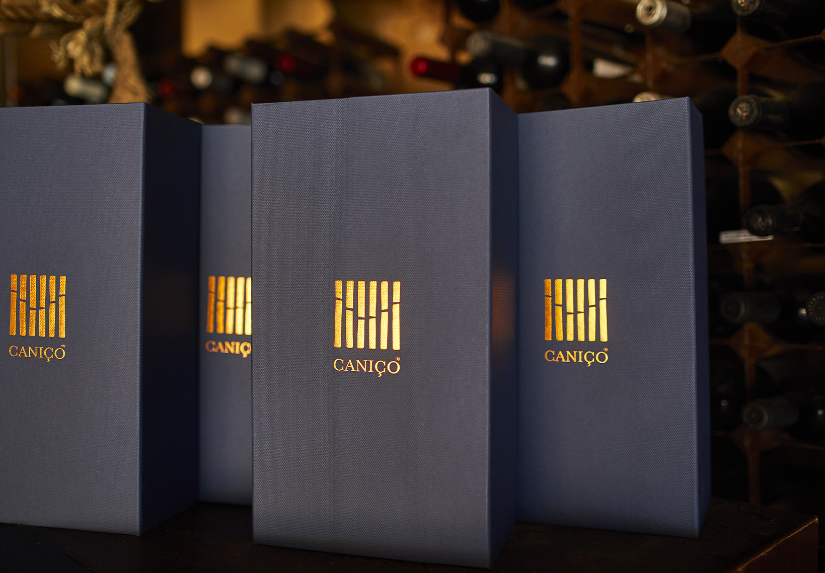 Três caixas azuis escuras com o logótipo dourado do Caniço, alinhadas em frente a uma garrafeira de vinhos.