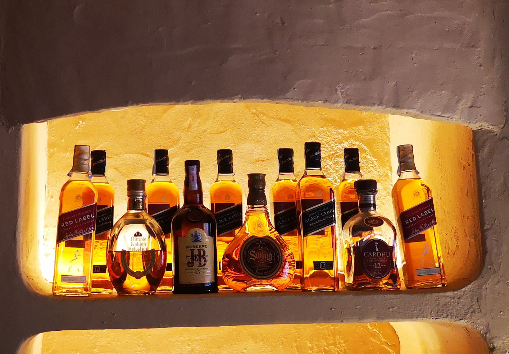 Diversas garrafas de whisky de marcas premium expostas num nicho de parede iluminado com luz quente.