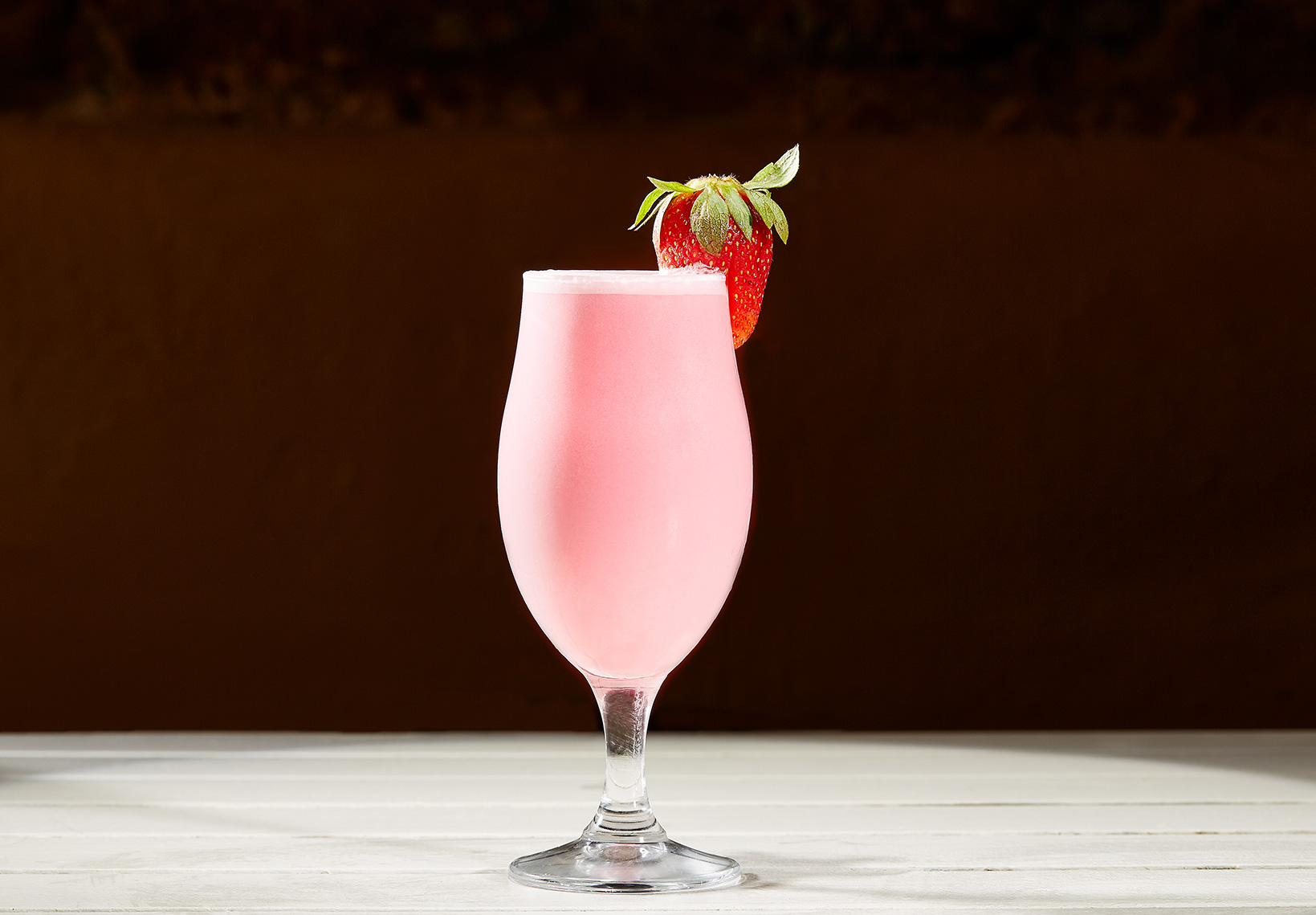 Um copo de pé alto com uma bebida cremosa cor-de-rosa, decorado com um morango fresco na borda.