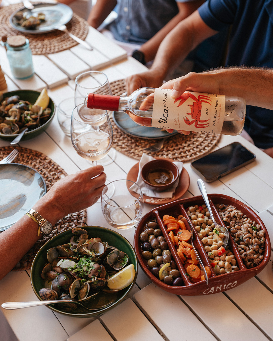 Pessoa a servir vinho branco numa mesa repleta de petiscos como amêijoas e saladas de leguminosas.