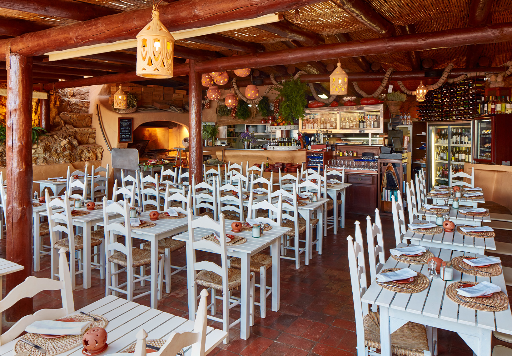 Interior do restaurante com mesas de madeira branca, cadeiras de palha e teto rústico de canas.
