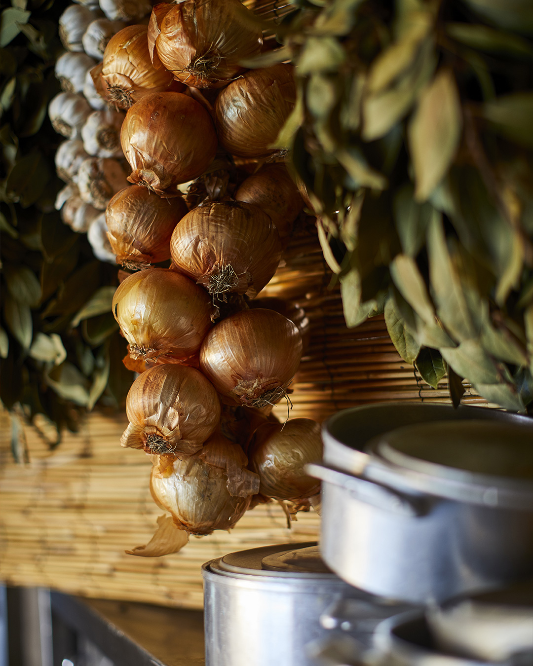 Pormenor de uma trança de cebolas douradas, alhos e ramos de loureiro secos suspensos numa parede de canas. / Close-up of a braid of golden onions, garlic, and dried bay leaf branches hanging against a bamboo reed wall.