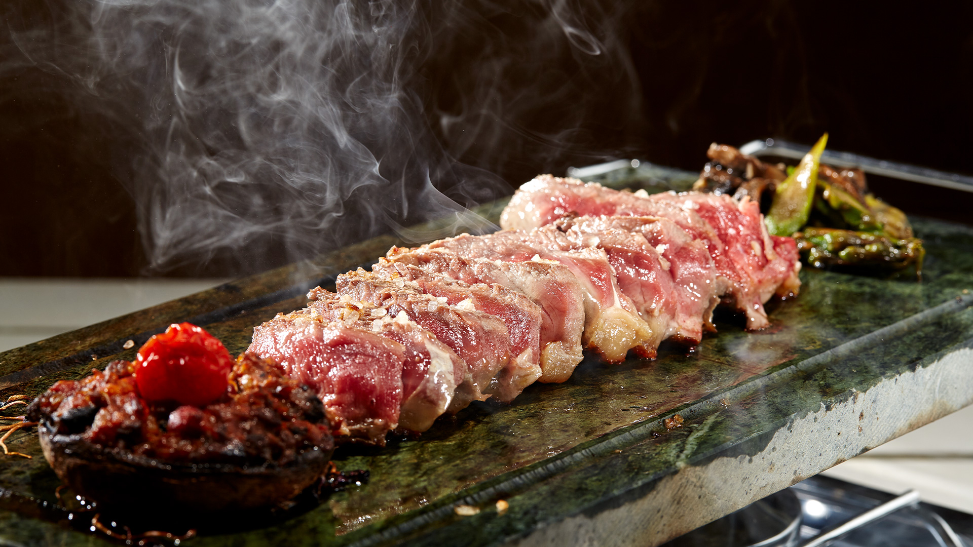 Carne fatiada sobre uma pedra quente com fumo a sair, acompanhada por legumes.