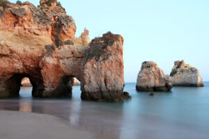 Paisagem costeira com falésias de tons ocres e arcos naturais sobre a areia da praia.